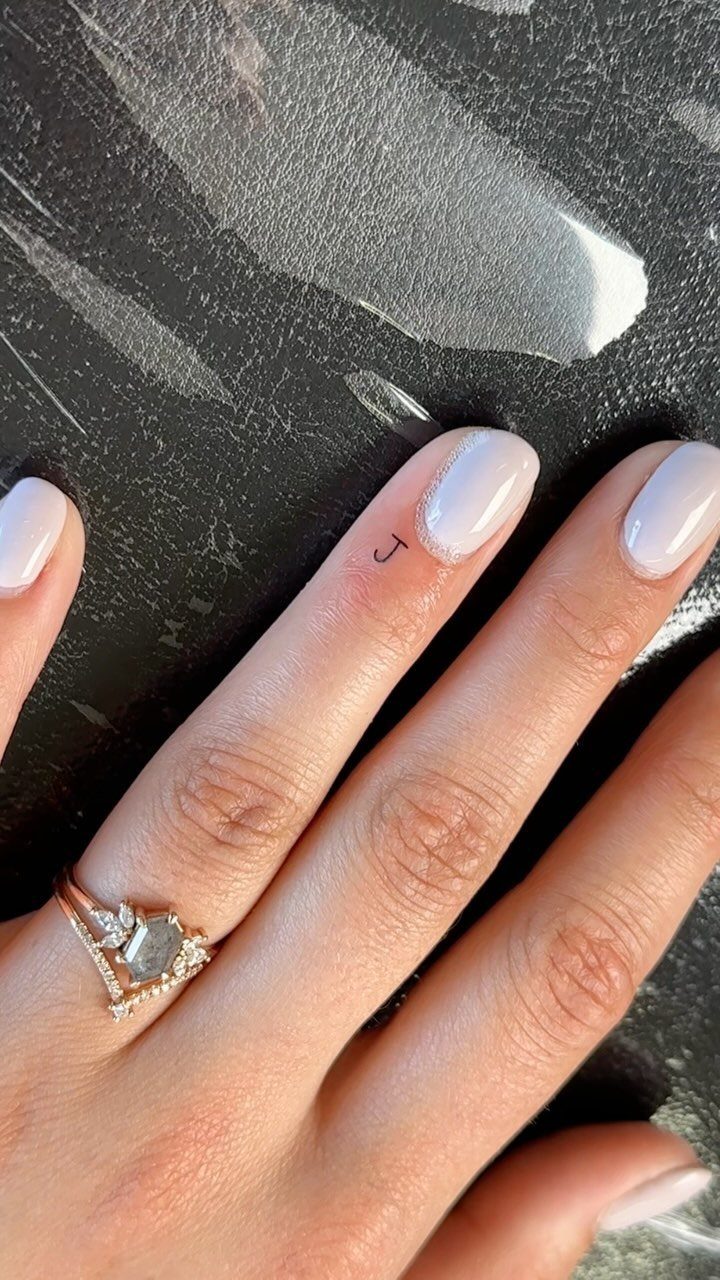 Tiny black letter J tattoo on a finger with white nail polish.