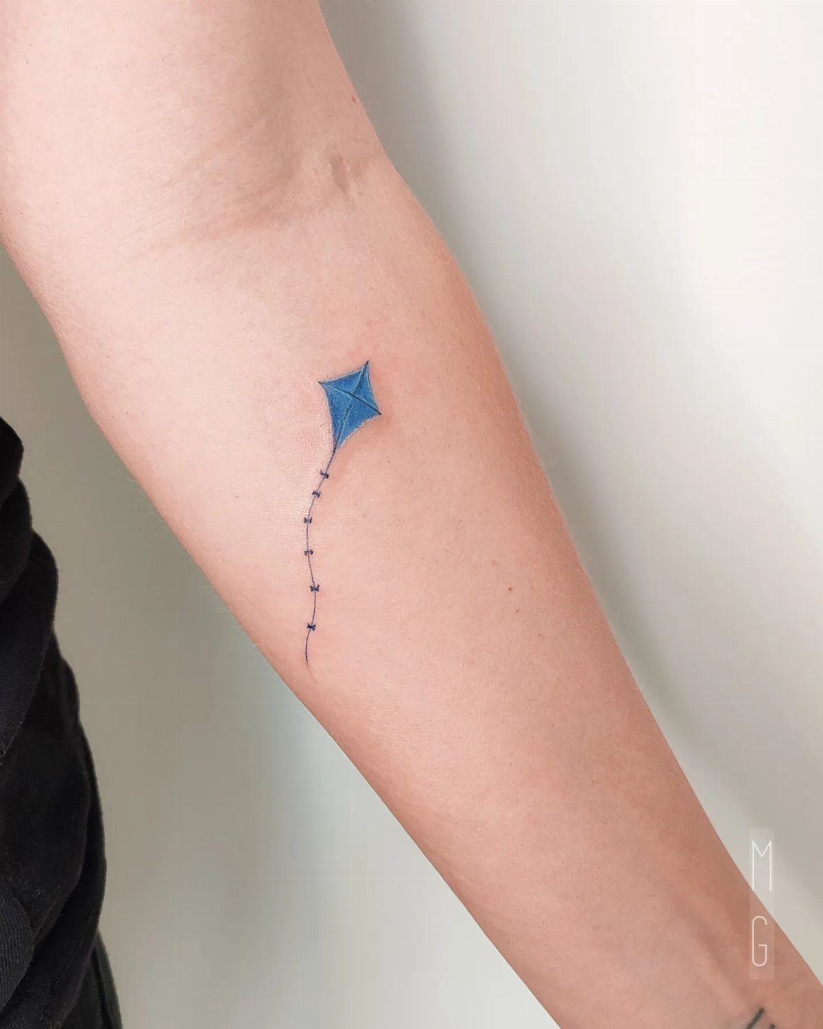 Small blue kite tattoo with a string on a person's inner forearm.