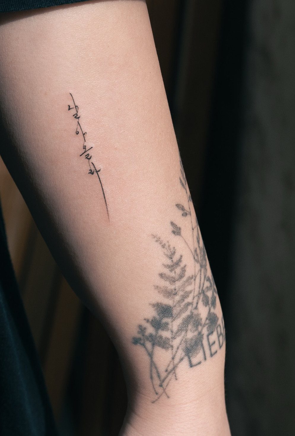 Forearm with a vertical character tattoo and a botanical design featuring the word LIEBE.