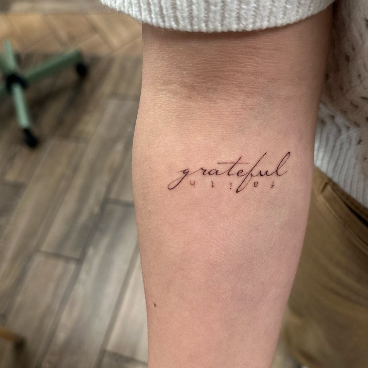 Forearm with a delicate cursive tattoo of the word grateful and faith.