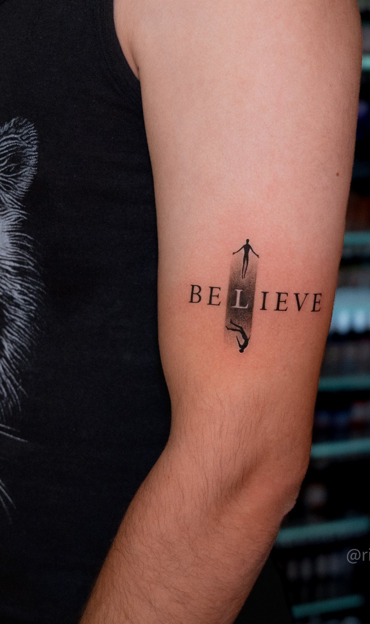 Tattoo on an arm showing the word BELIEVE with surreal silhouette figures inside.