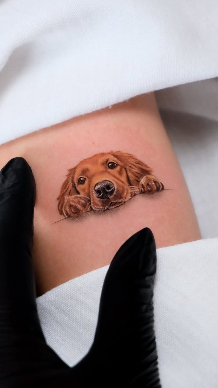 Small realistic golden retriever tattoo on skin with a gloved hand nearby.