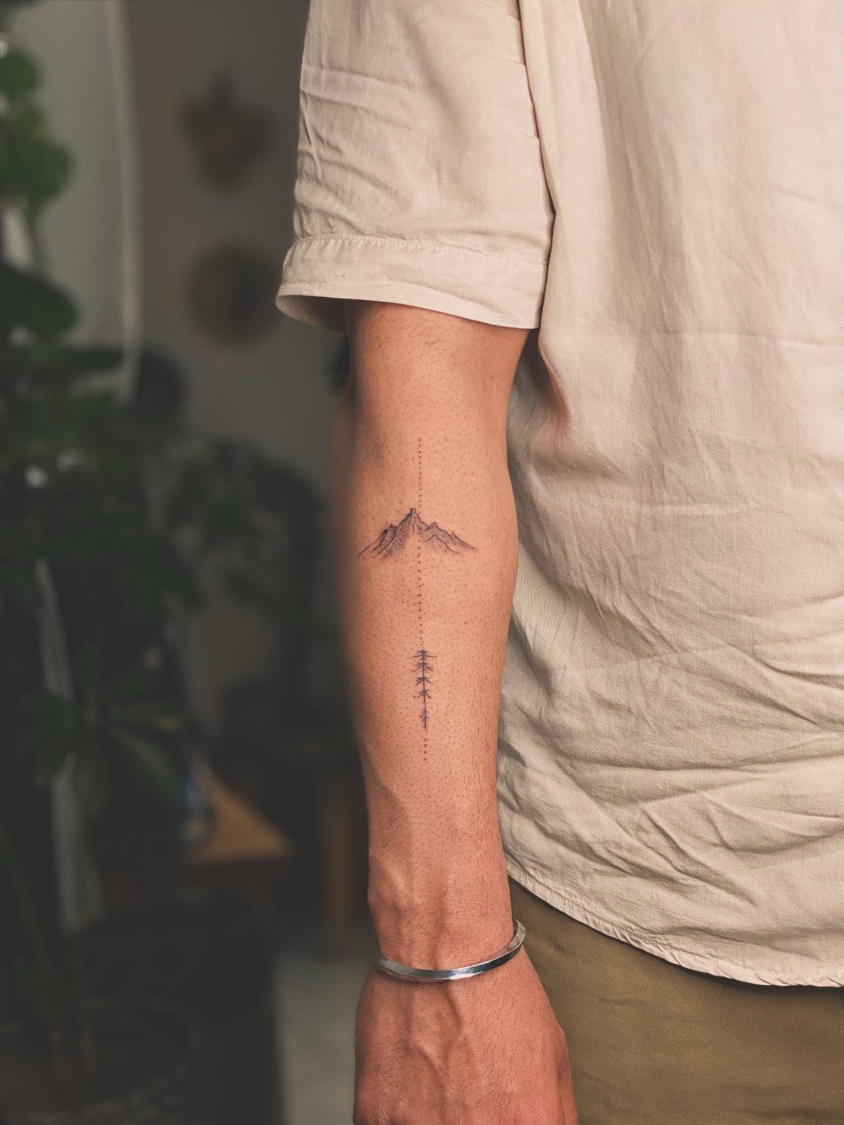 Minimalist mountain tattoo on a forearm with a vertical line and small tree.