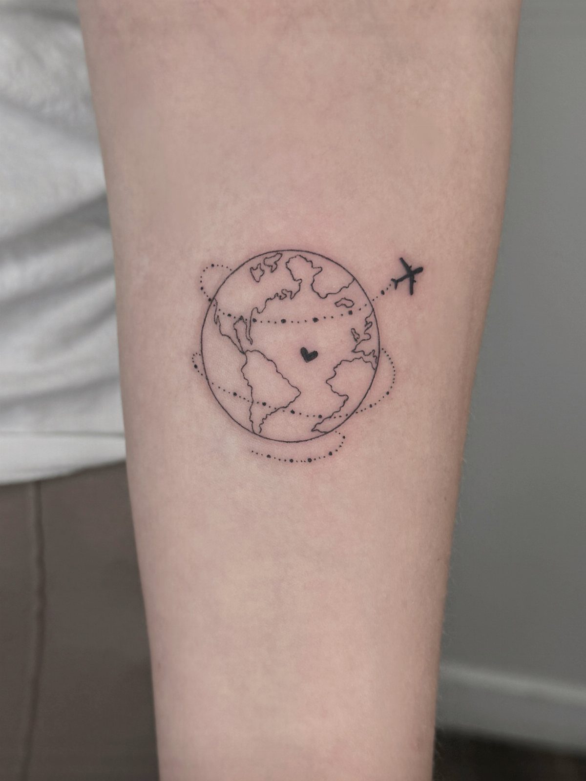 Minimalist globe tattoo with a small heart and airplane on a forearm