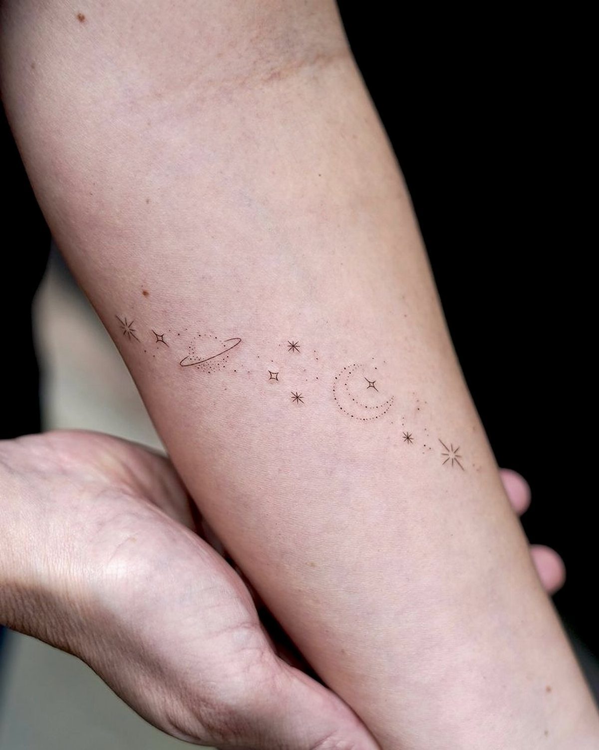 Fine line tattoo of a moon, planet, and stars on a forearm.