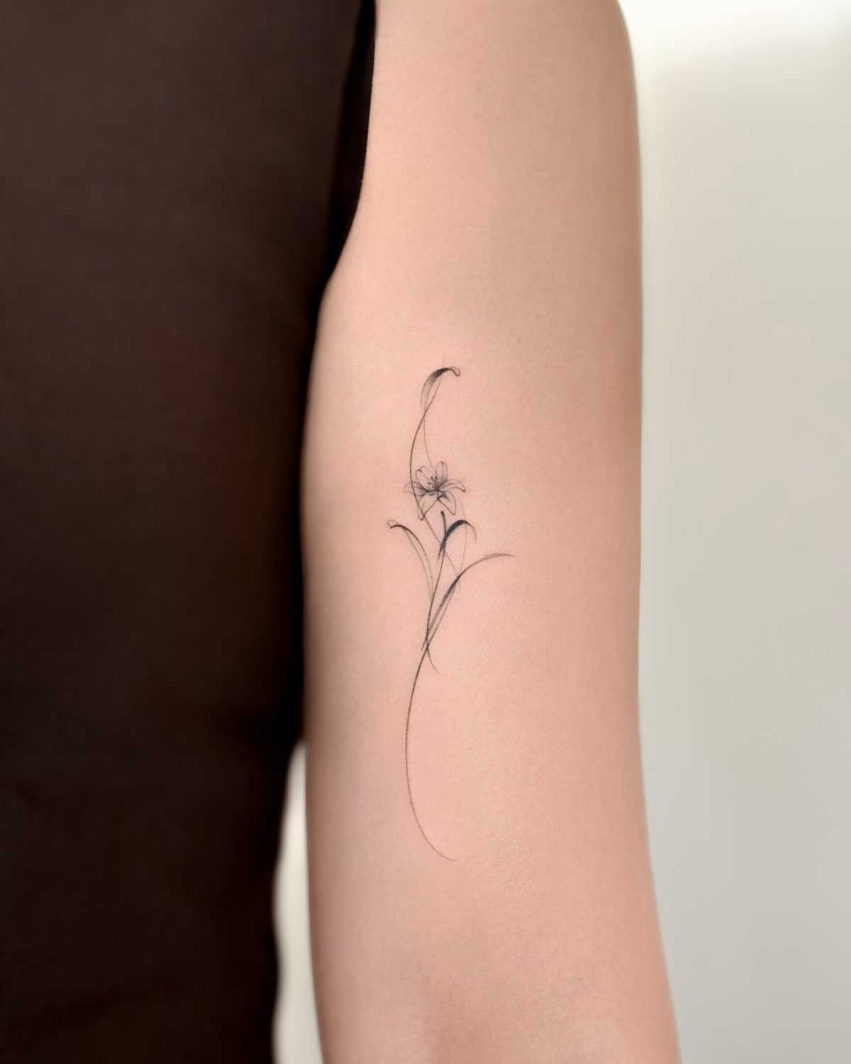 Fine line flower tattoo with a delicate stem on a person's upper arm.