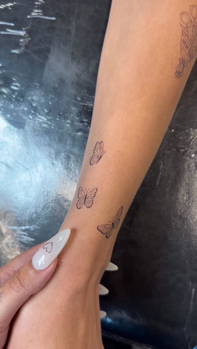 Three small fine line butterfly tattoos on a person's forearm with white nails.