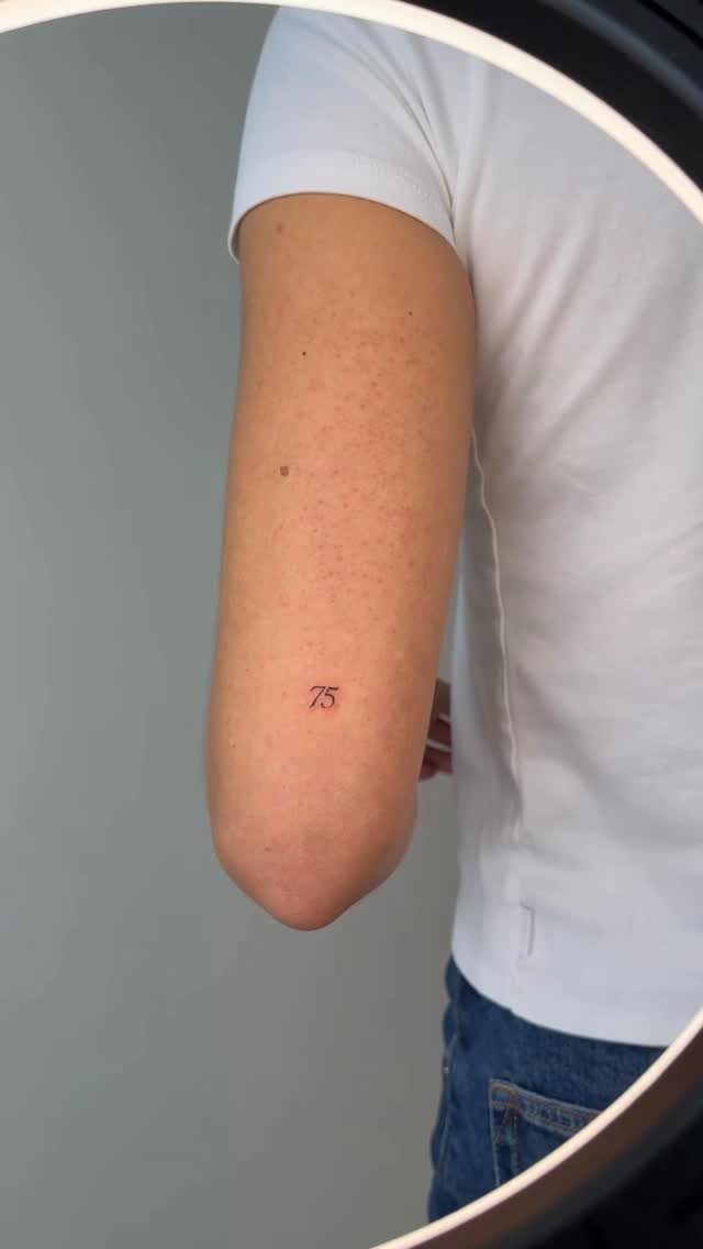 Small red fine line tattoo of number 75 on a person's upper arm.
