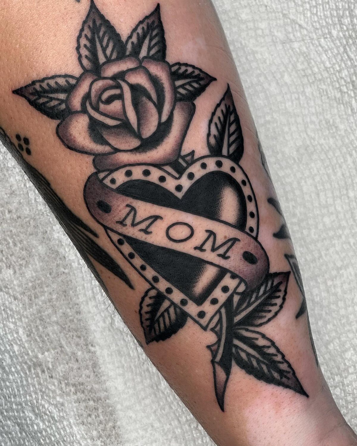Black and gray heart tattoo with Mom banner, rose, and arrow on skin