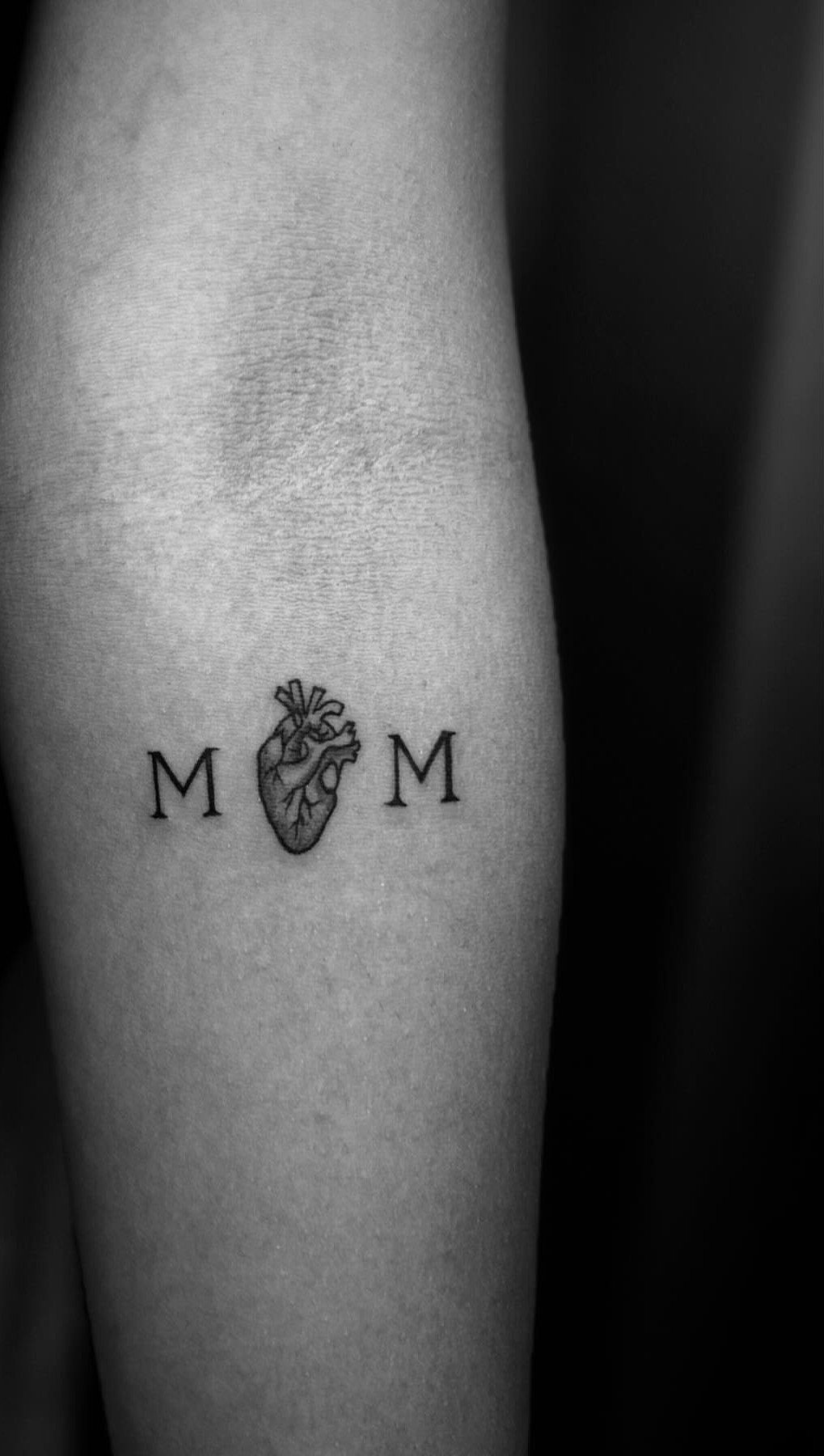 Anatomical heart tattoo spelling MOM on a person's forearm in black ink.