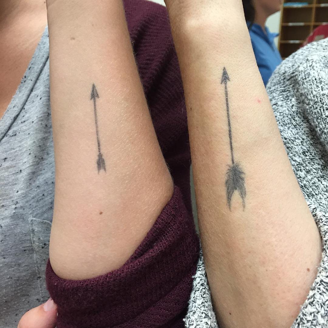 Two forearms with matching black arrow tattoos pointing upwards on the skin.