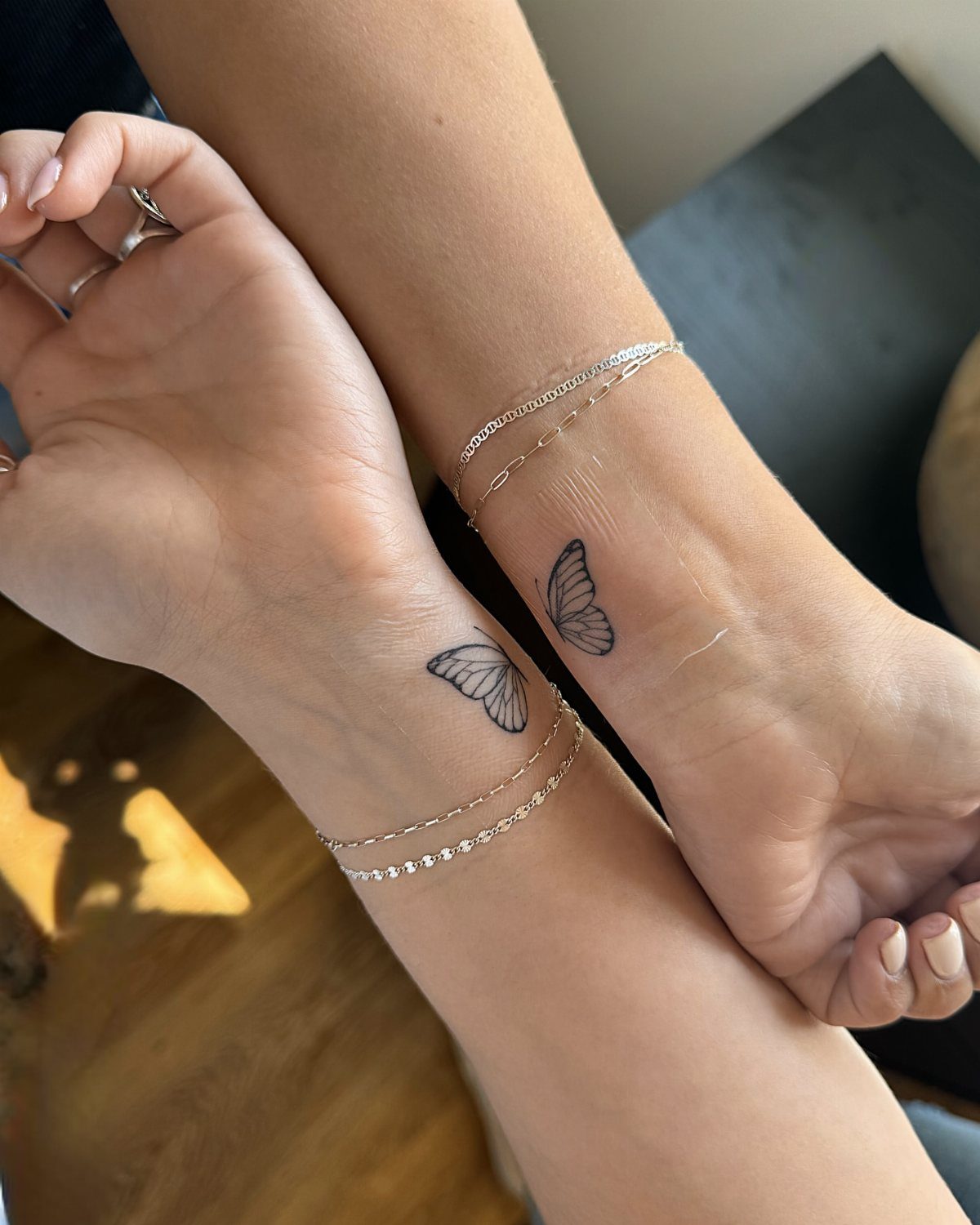 Two wrists with matching small black butterfly tattoos and silver chain bracelets.