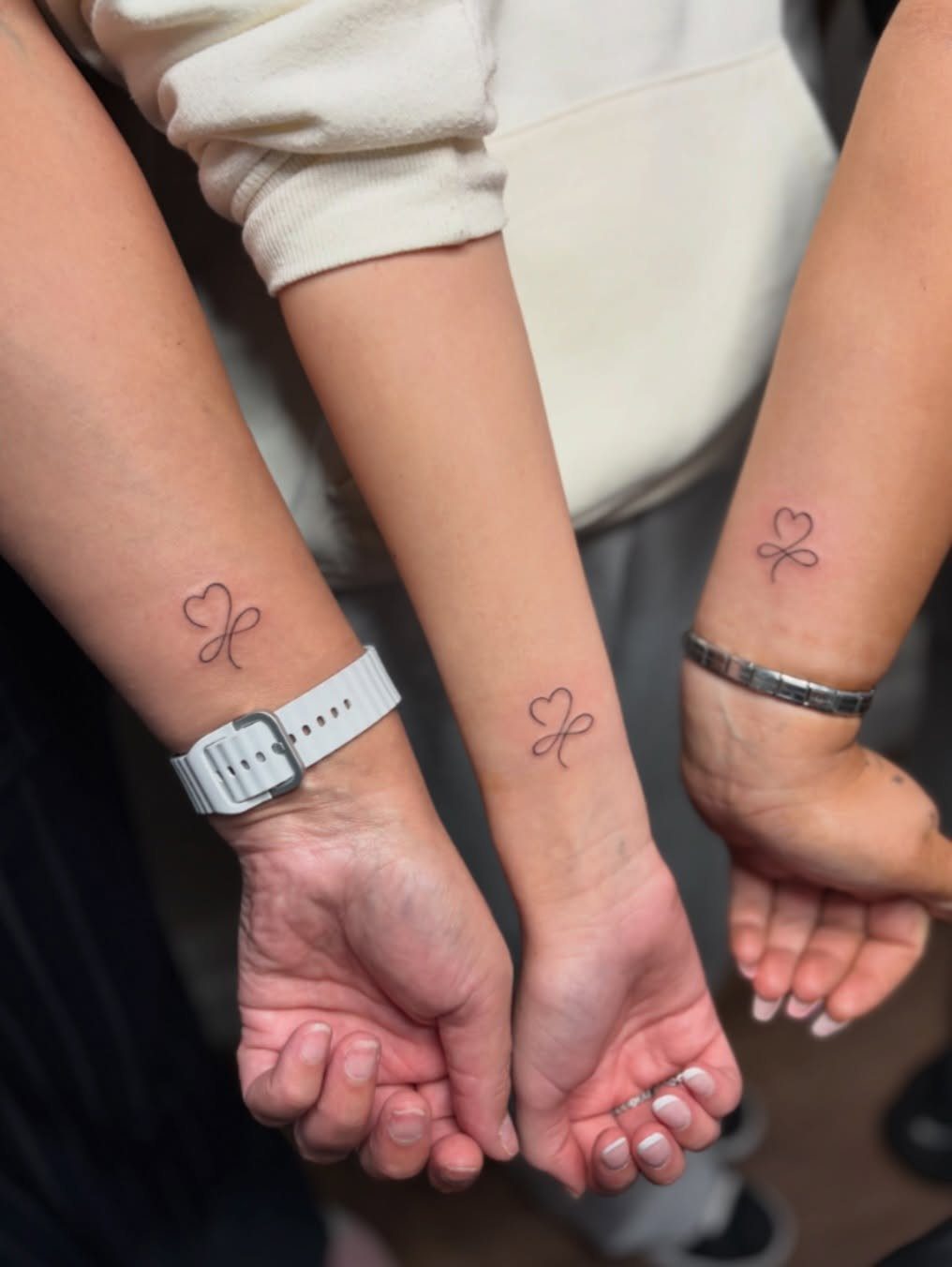 Three wrists showing matching small black heart knot tattoos in a row.