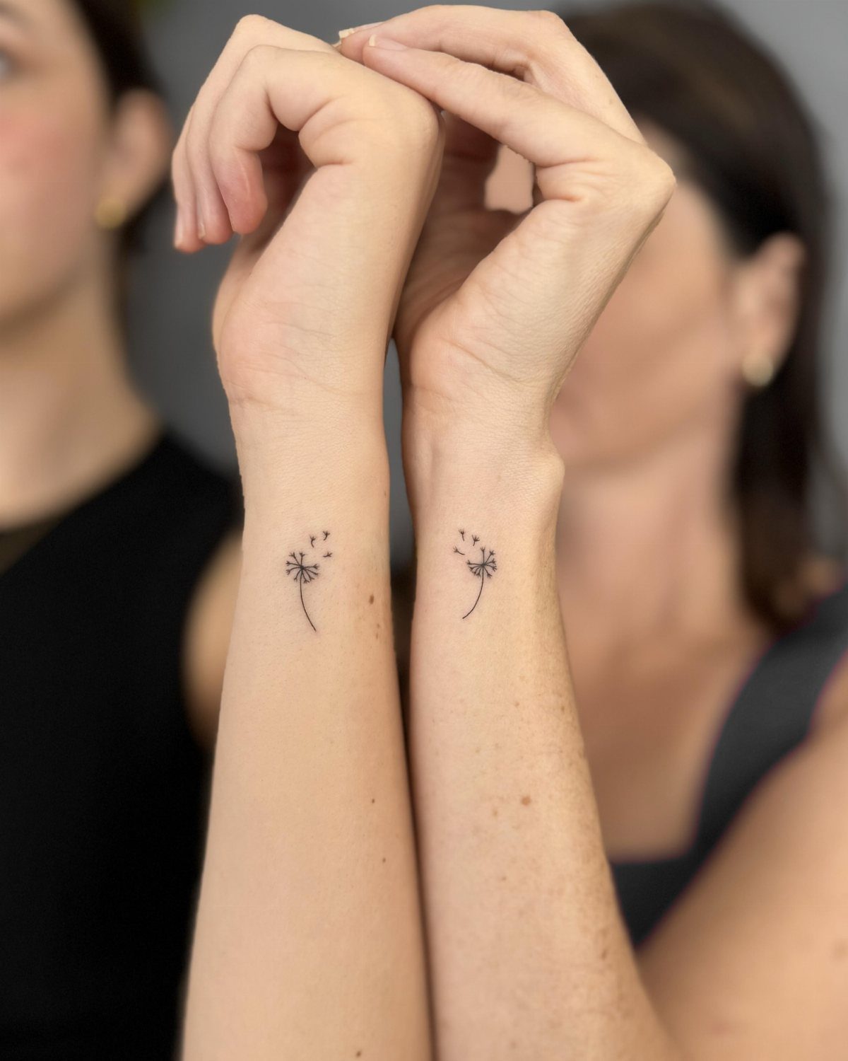 Two inner wrists with matching minimalist black dandelion tattoos held side by side.