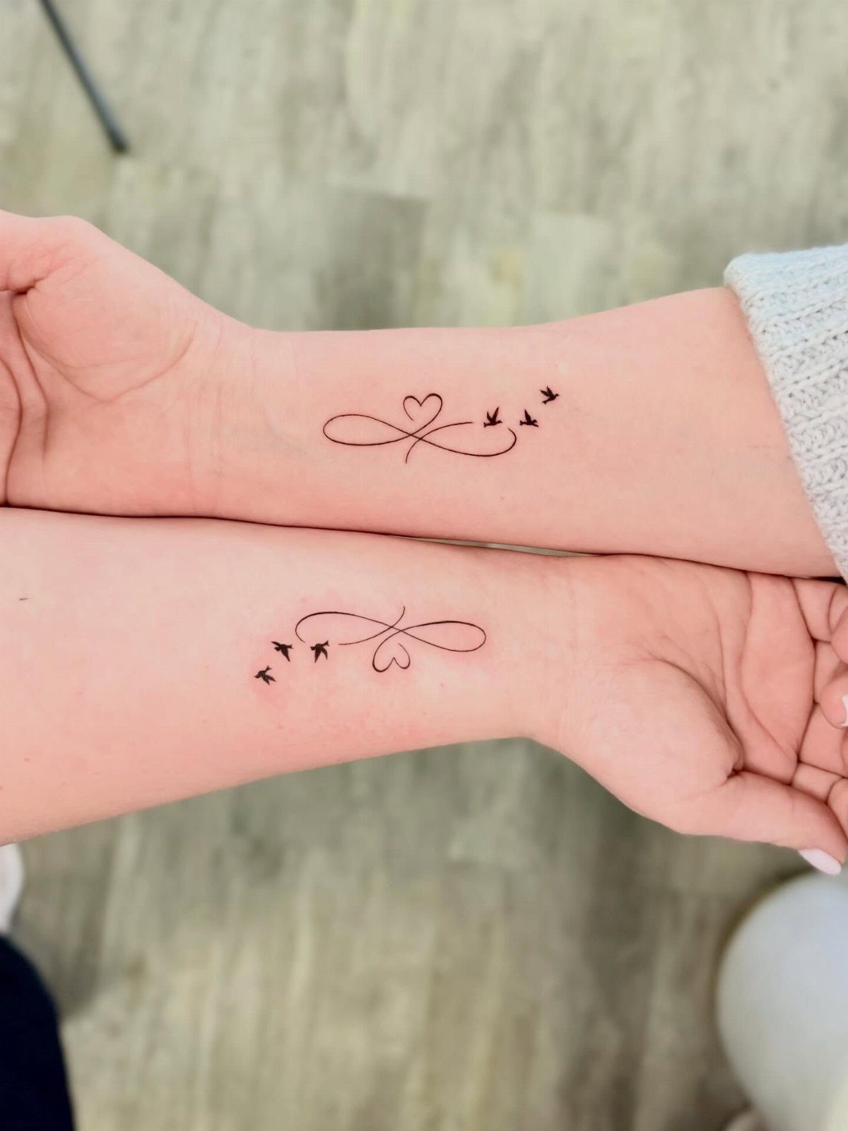 Matching infinity symbol tattoos with hearts and birds on two wrists.