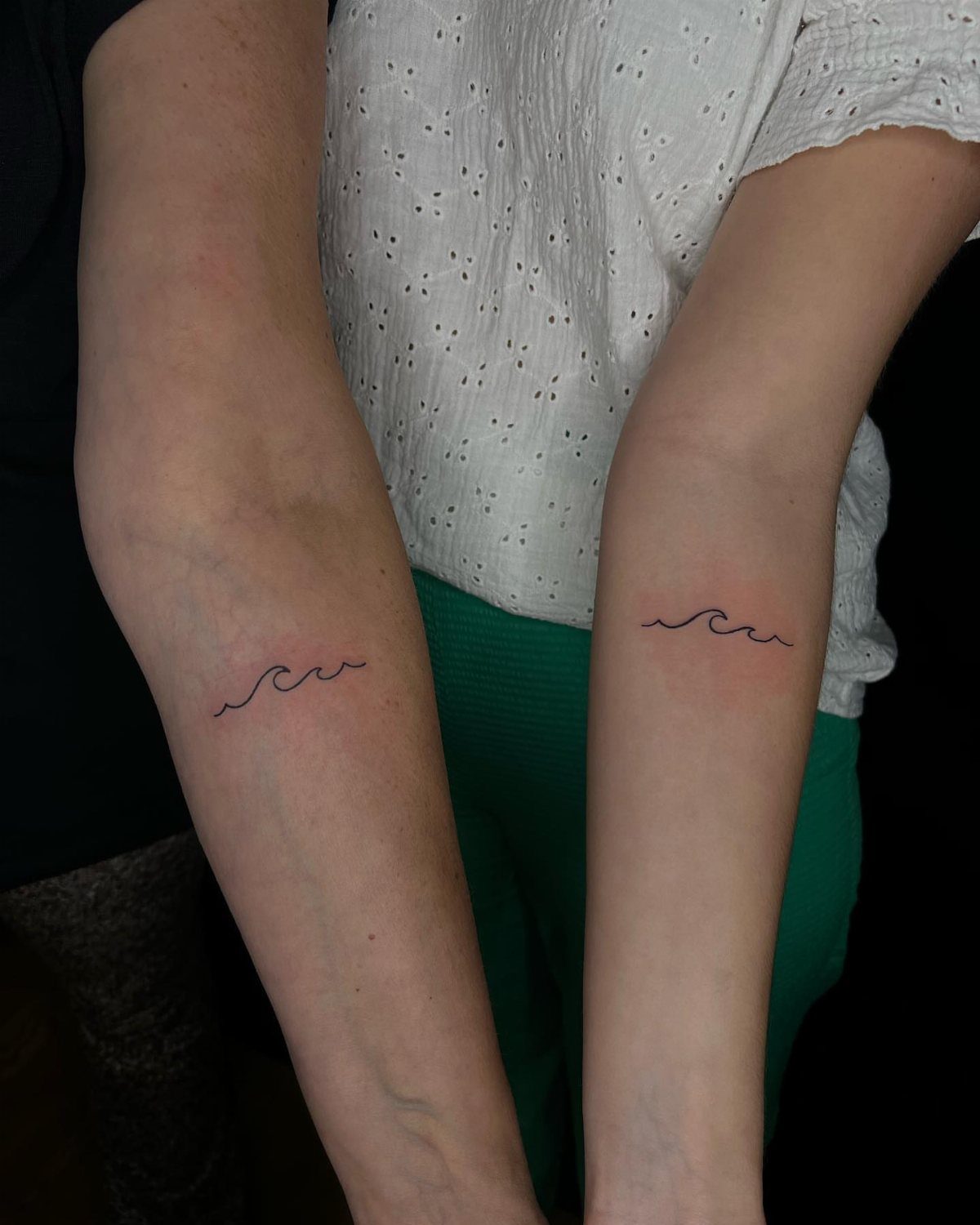 Two forearms with matching minimalist black ink ocean wave tattoos side by side.