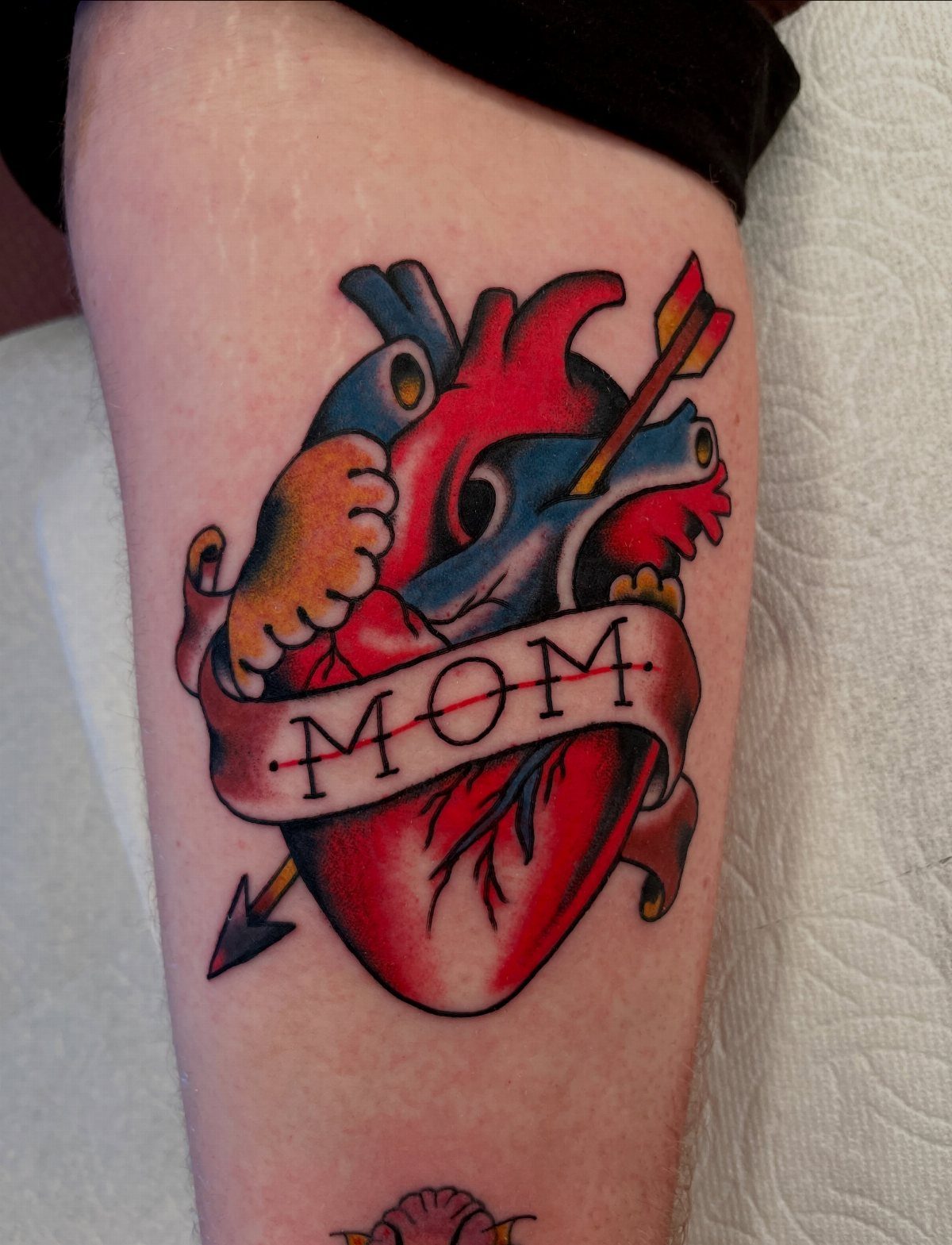 Anatomical heart tattoo with an arrow and a banner reading MOM on skin.