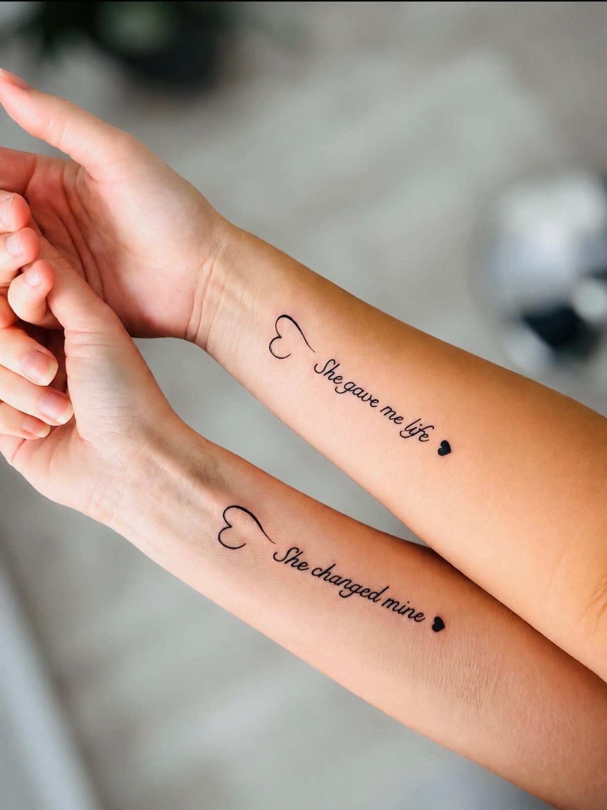 Two forearms with minimalist heart tattoos and cursive text about a mother.
