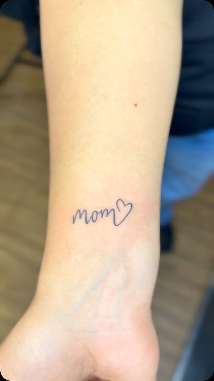 Minimalist mom heart tattoo in black ink on a person's inner wrist