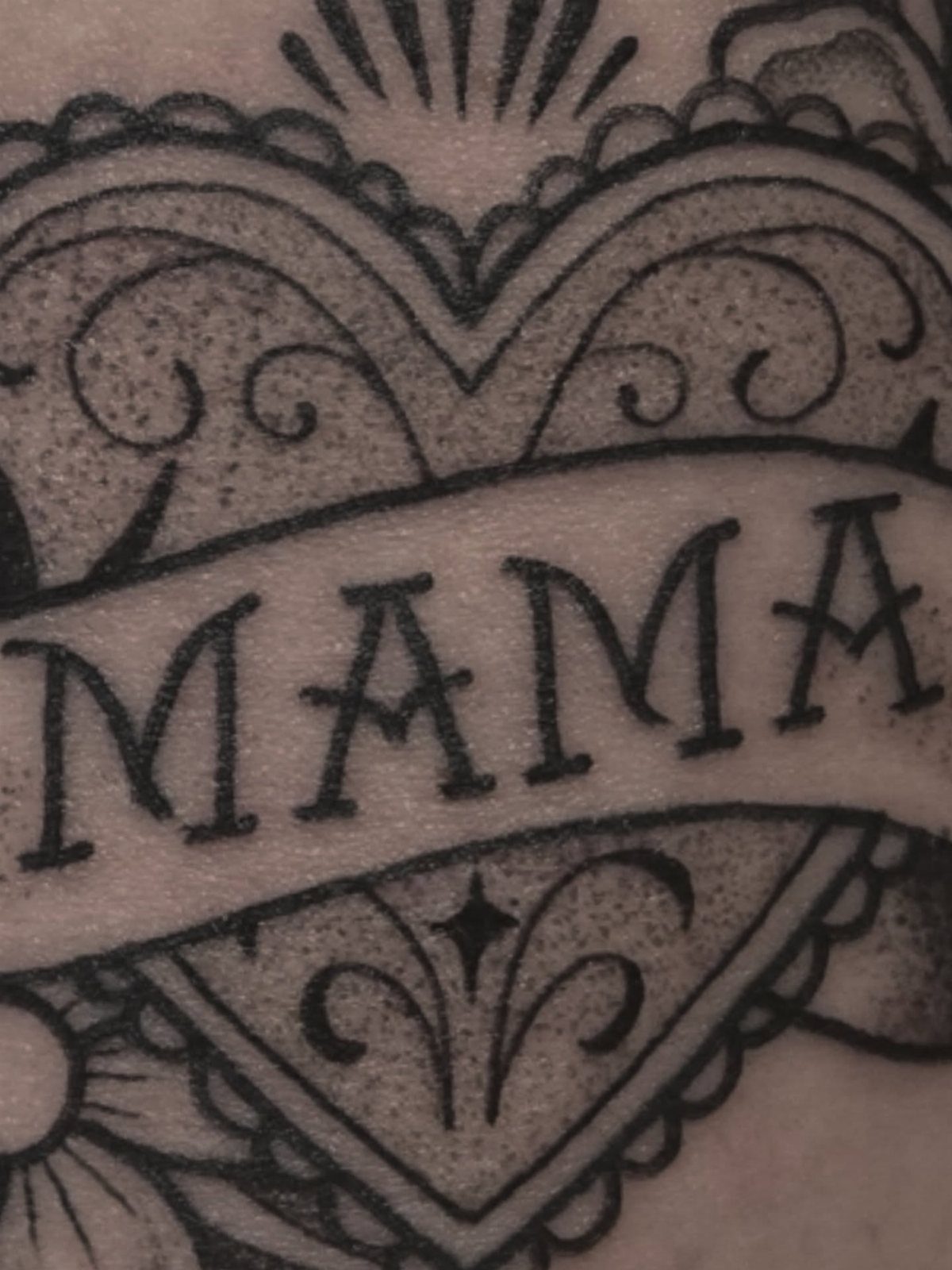 Heart tattoo with a banner reading MAMA and ornate decorative swirls on skin.