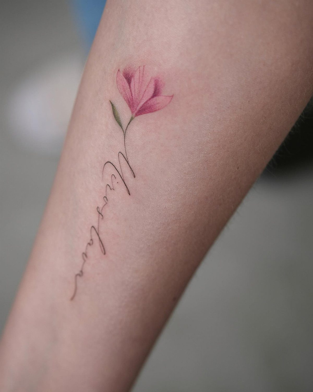 Cursive name tattoo on a forearm topped with a pink flower blossom