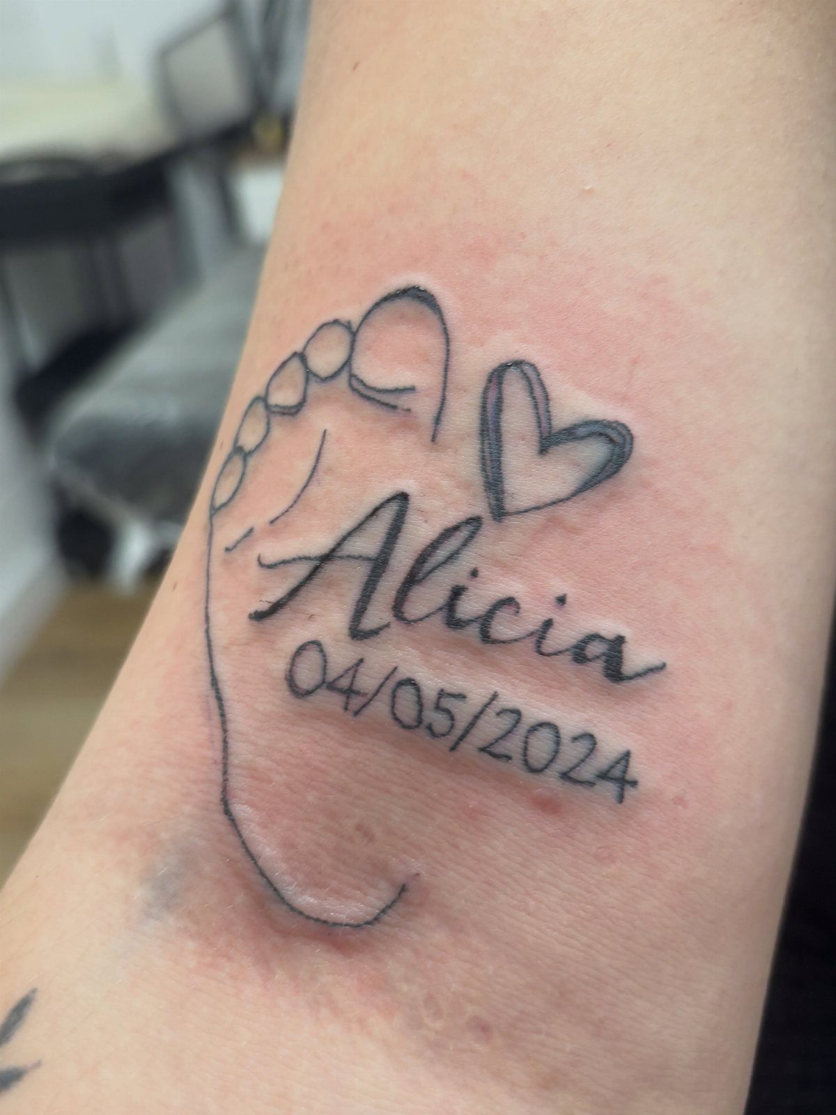 Tattoo of a baby footprint with the name Alicia and a date.