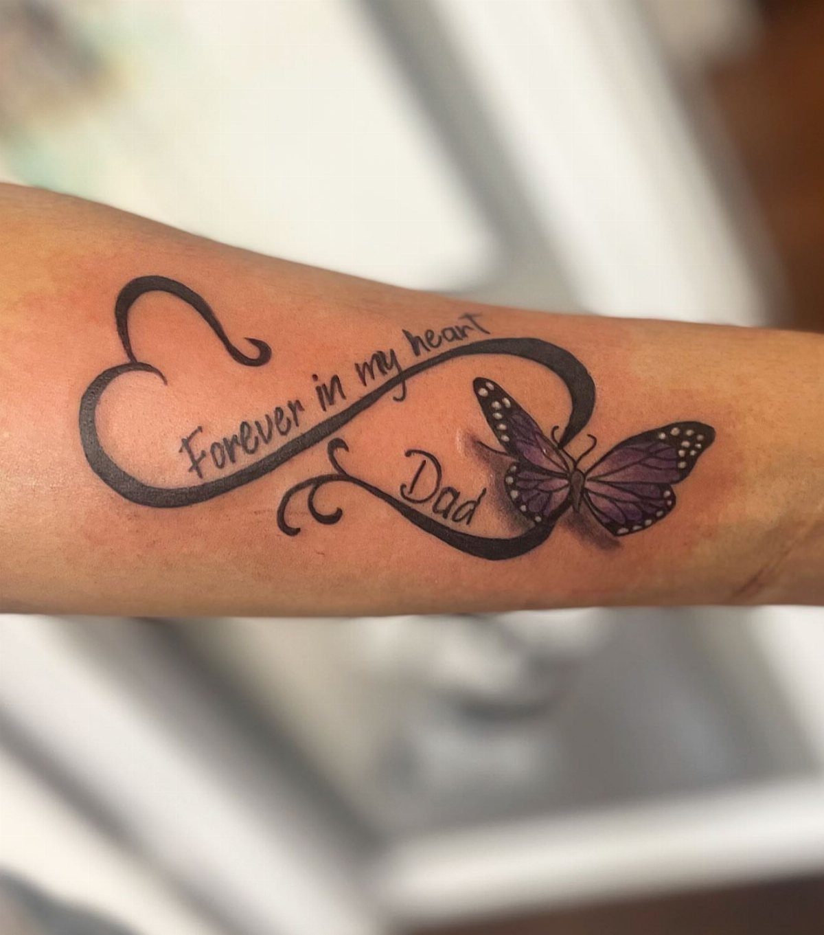 Forearm tattoo with infinity symbol, purple butterfly, and text reading Forever in my heart Dad