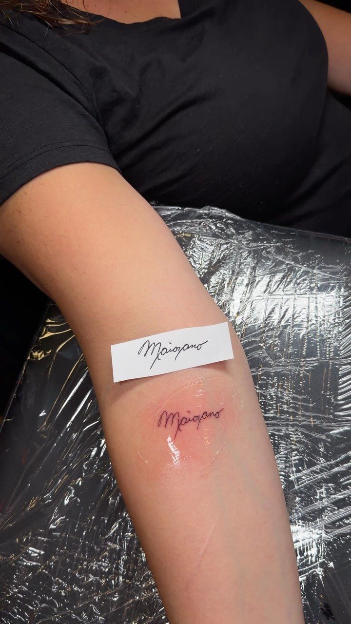 Handwritten name tattoo in red ink on a forearm with reference paper