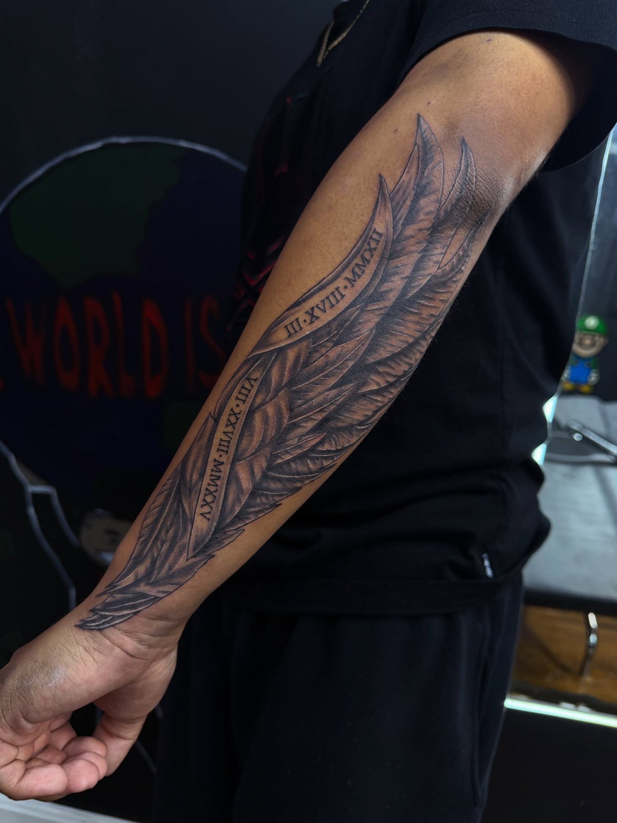 Forearm tattoo of a detailed feather wing containing integrated Roman numeral dates.
