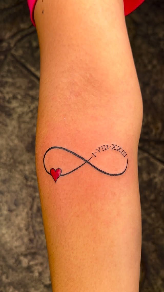 Infinity symbol tattoo with Roman numerals and a small red heart on a forearm