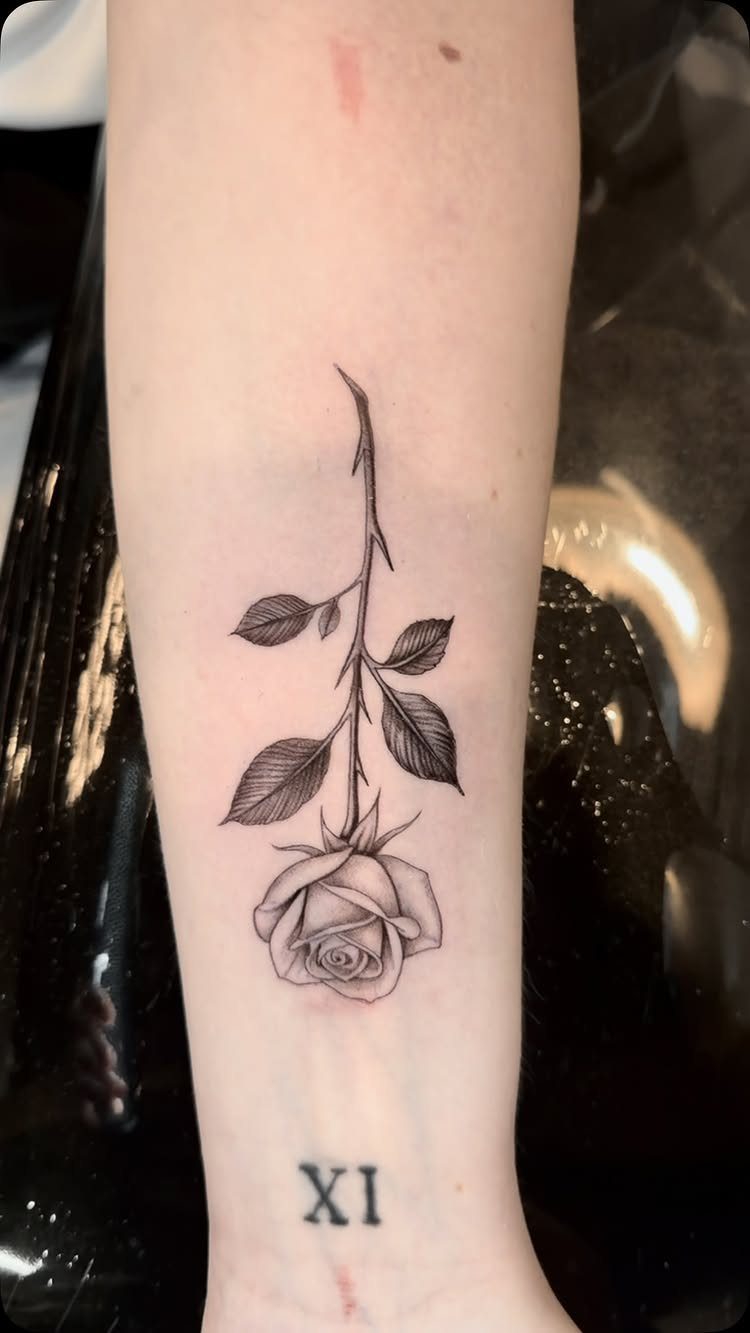 Forearm with a detailed rose tattoo and a small Roman numeral XI below.