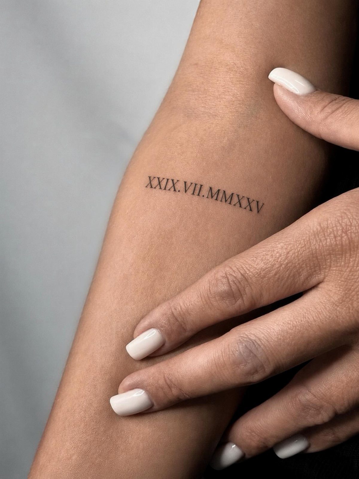 Small Roman numeral date tattoo on a person's forearm with white nails.