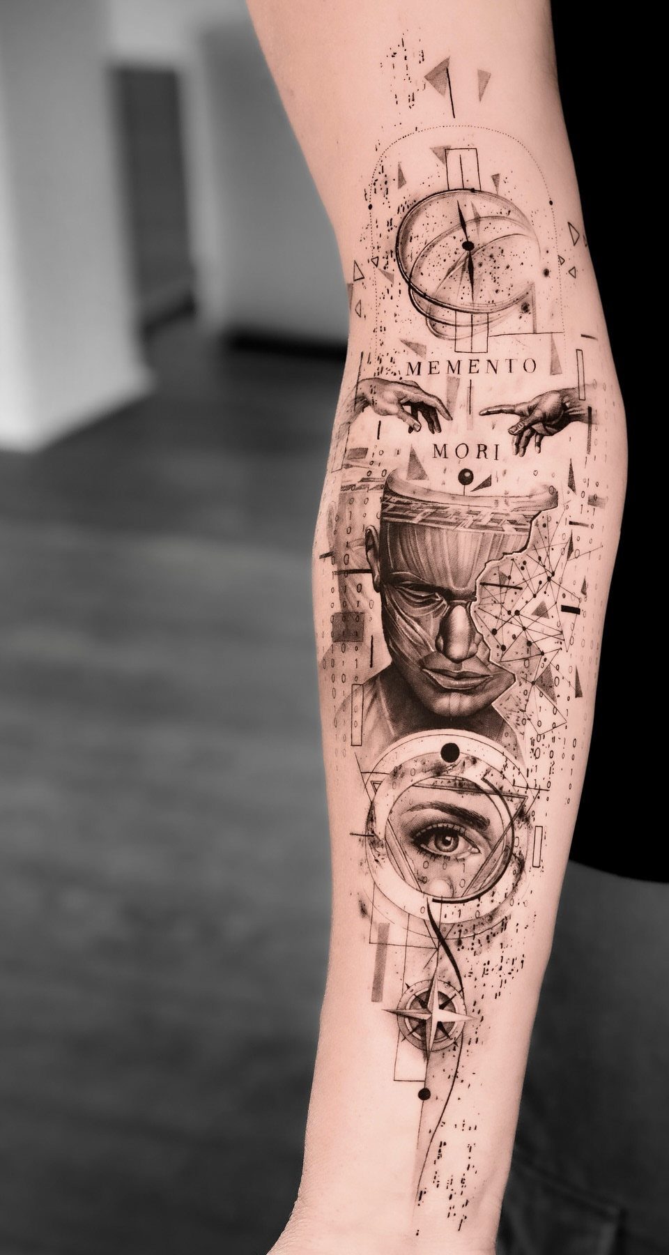 Surreal black and grey forearm tattoo featuring a face, eye, and geometric elements.