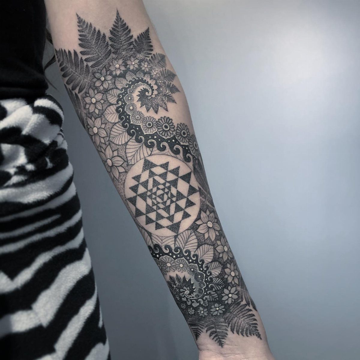 Forearm tattoo featuring a central sacred geometry star surrounded by floral and fern patterns.