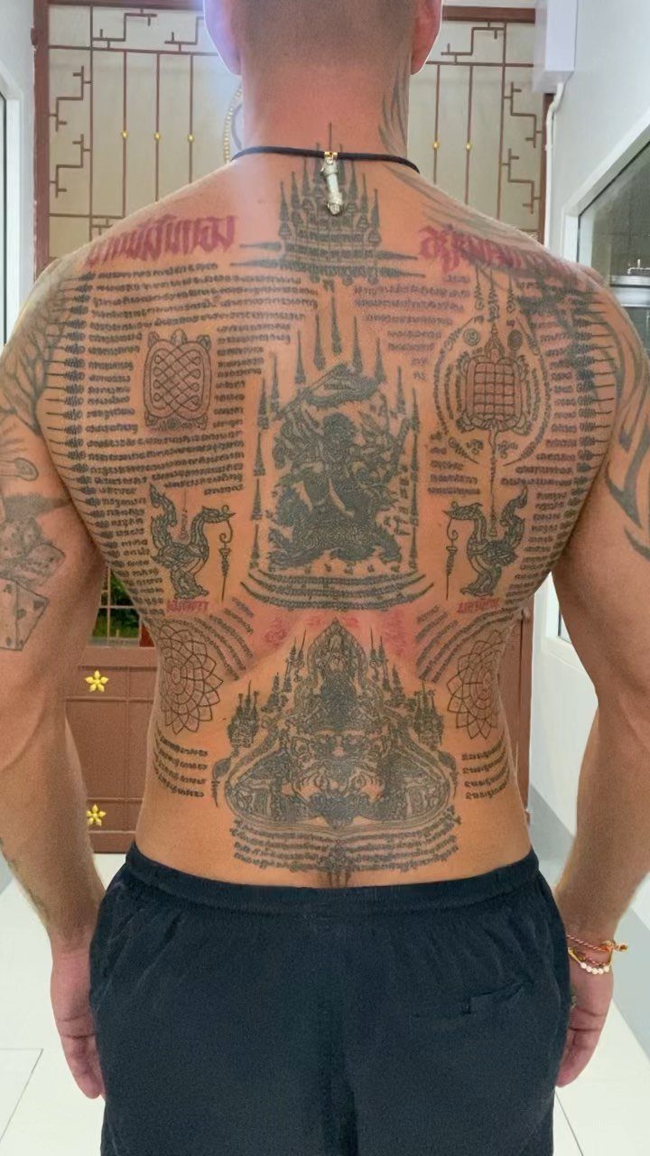 Man's back covered in intricate black ink Sak Yant tattoos with traditional Thai scripts.