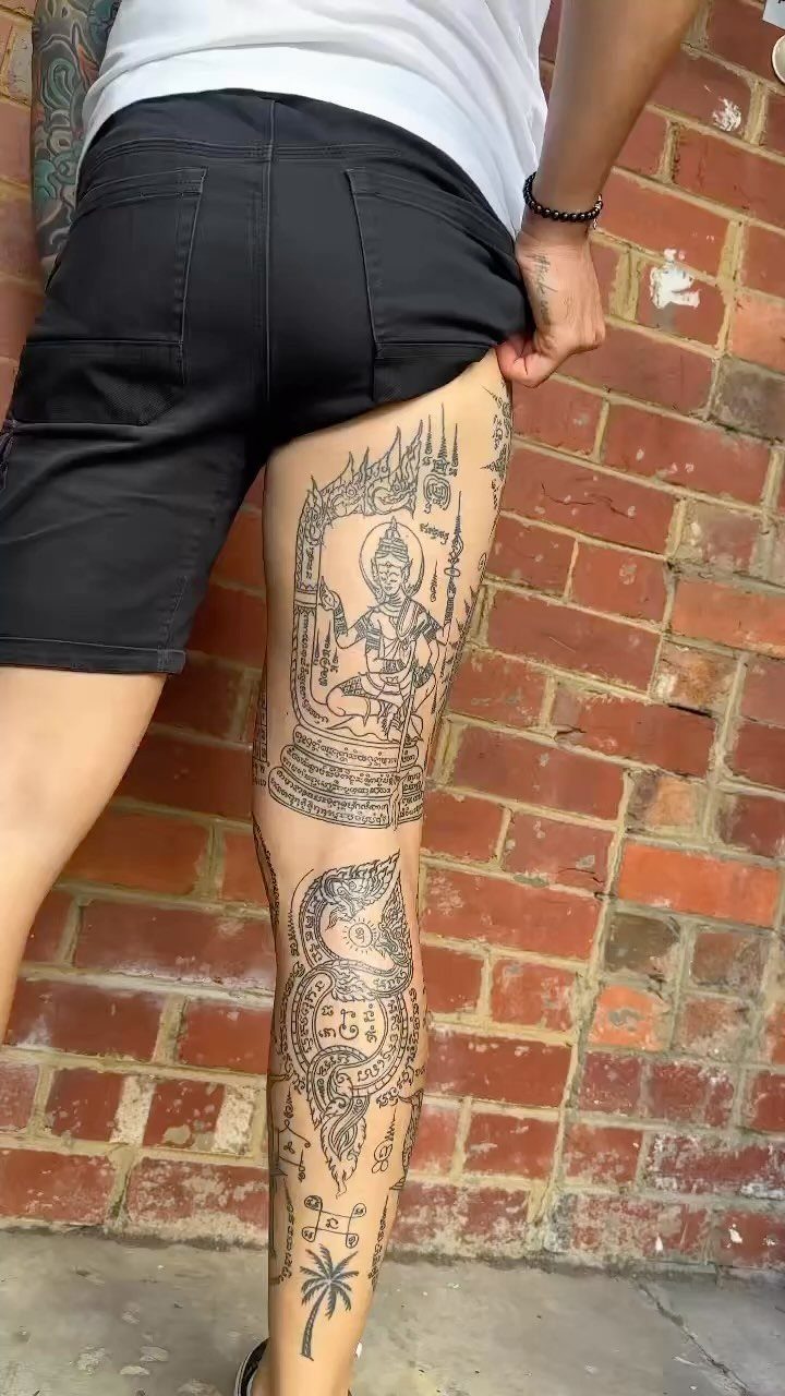 Detailed black ink Sak Yant tattoos on a person's leg against brick wall