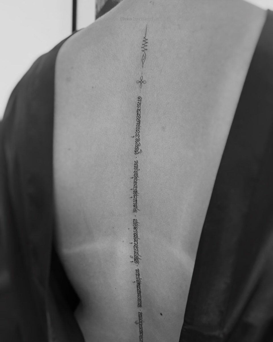 Vertical sak yant tattoo with Thai script on a person's spine in monochrome.