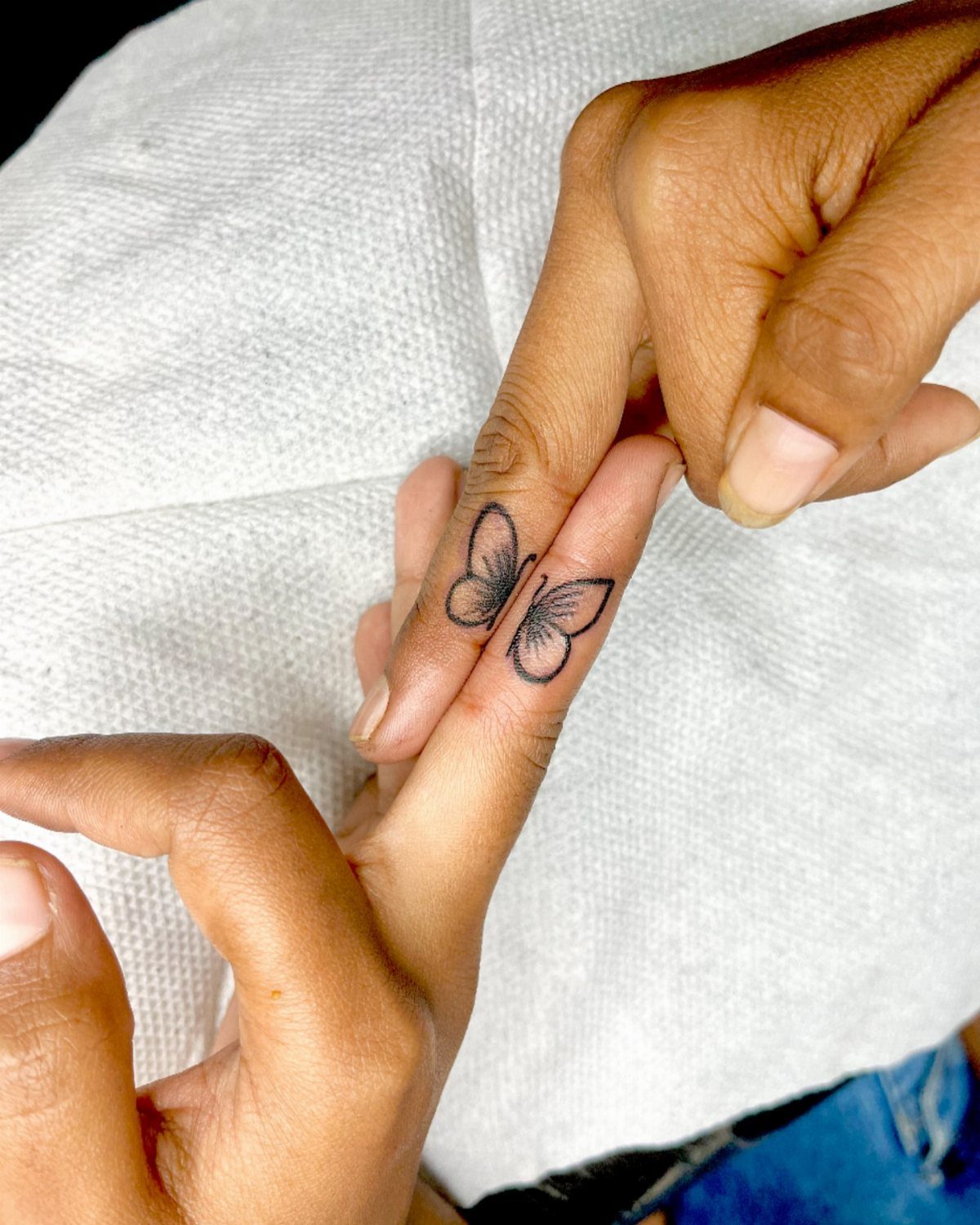 Two fingers with matching butterfly tattoos forming one complete butterfly on white fabric.