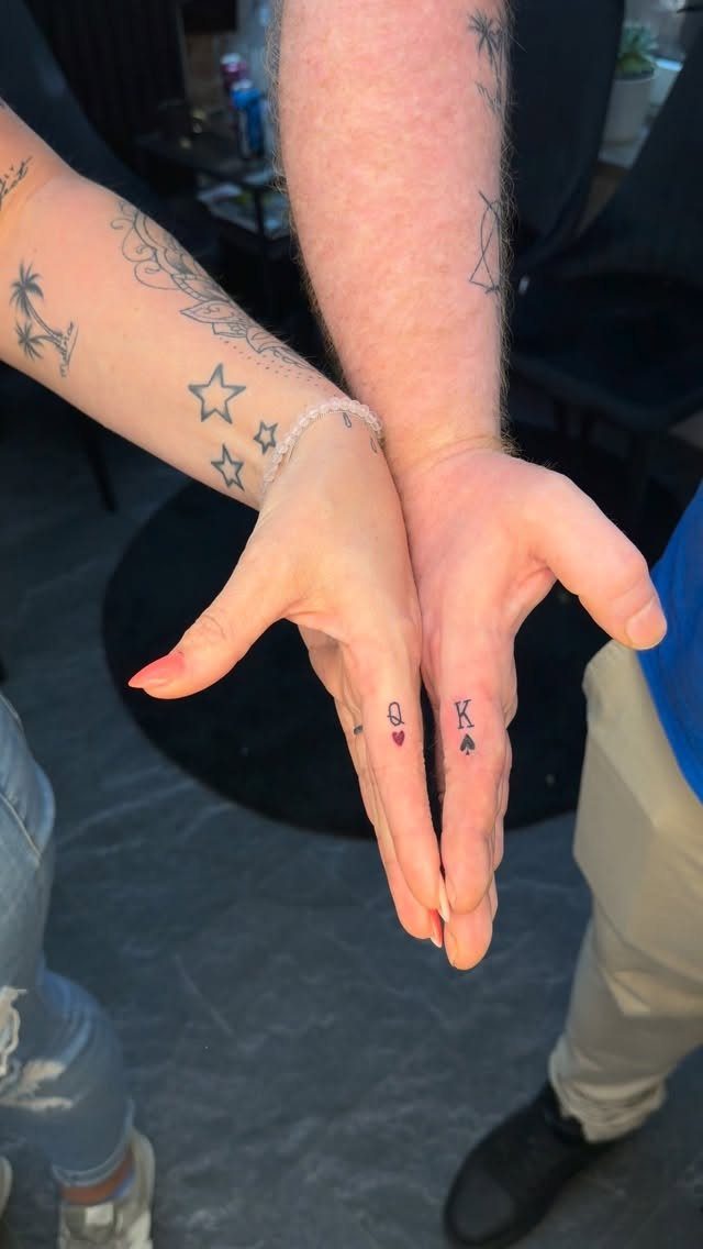 Two ring fingers with matching King and Queen tattoos featuring heart and spade symbols.