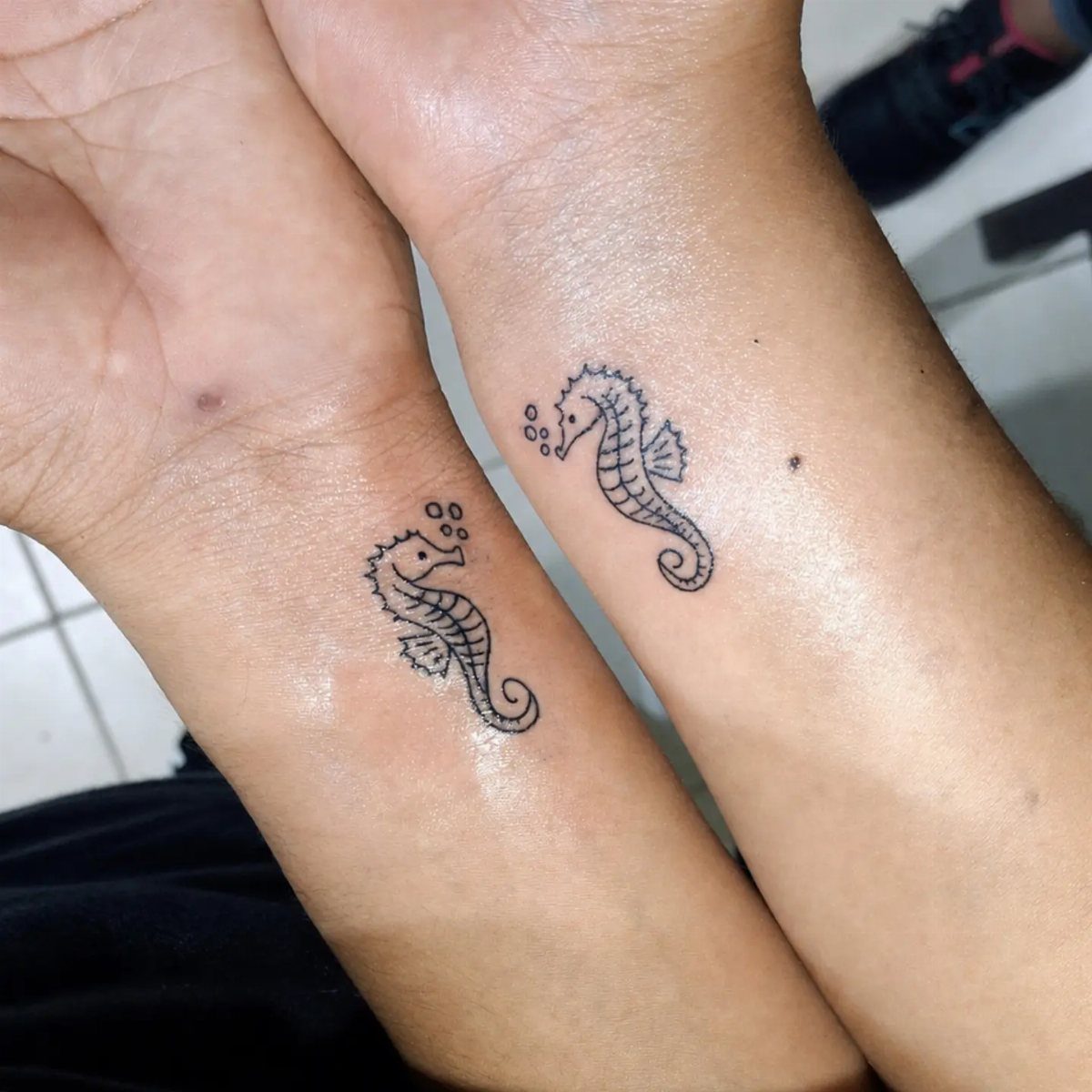 Matching seahorse tattoos on the wrists of two people against a tiled background.