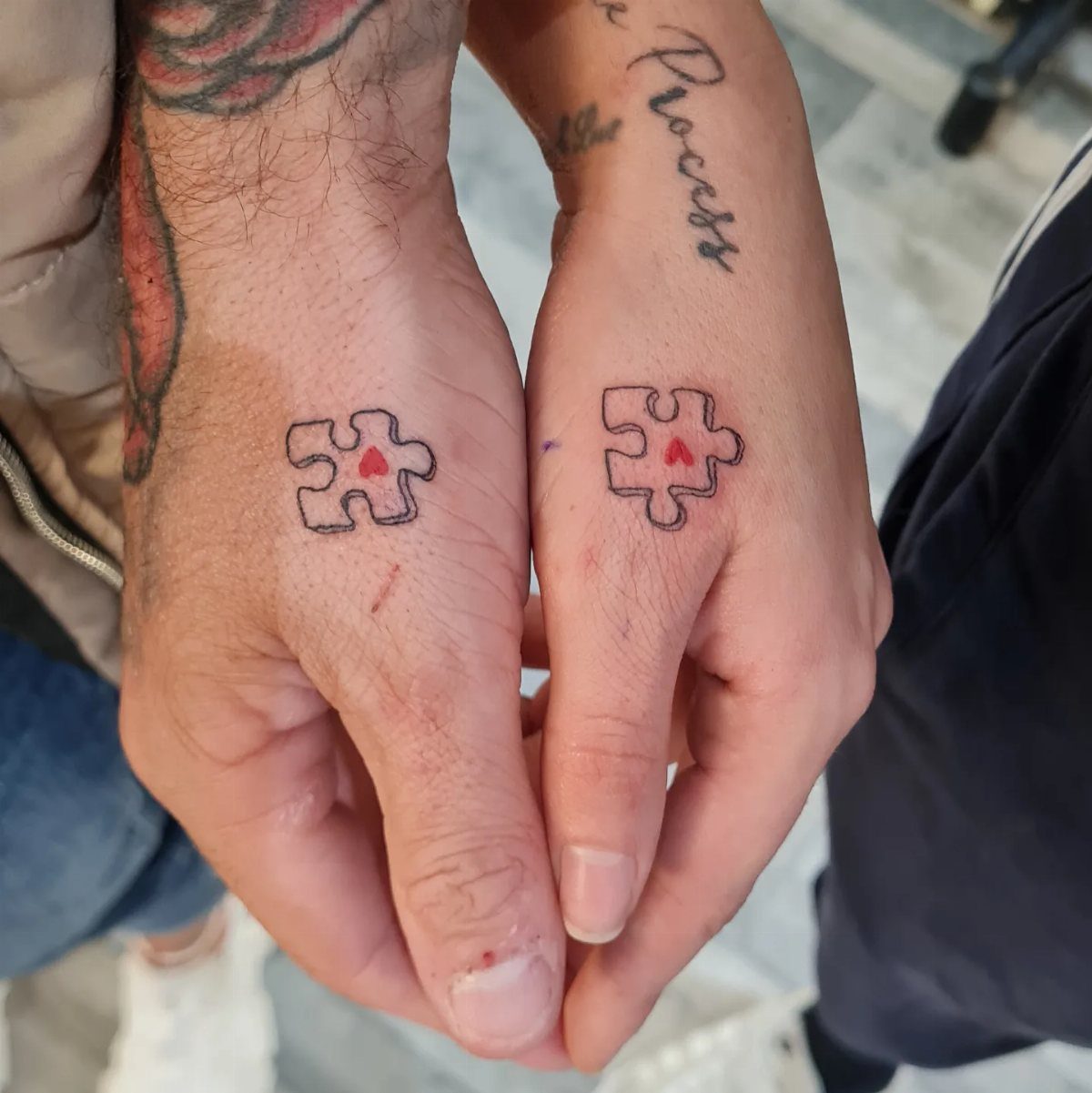 Two hands with matching puzzle piece tattoos containing small red hearts.