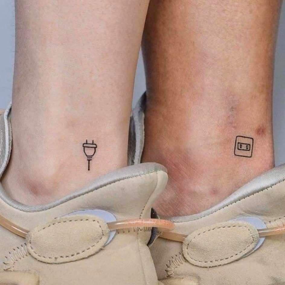 Two ankles with matching small tattoos of an electrical plug and socket.