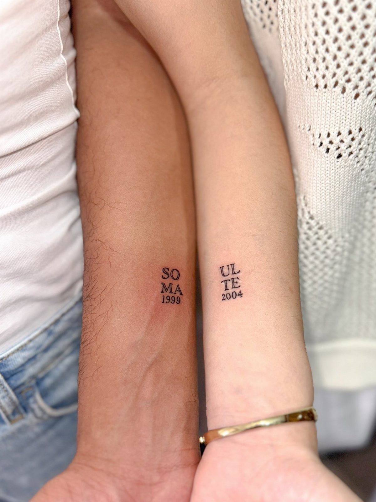 Two inner wrists with matching small black text and year tattoos.