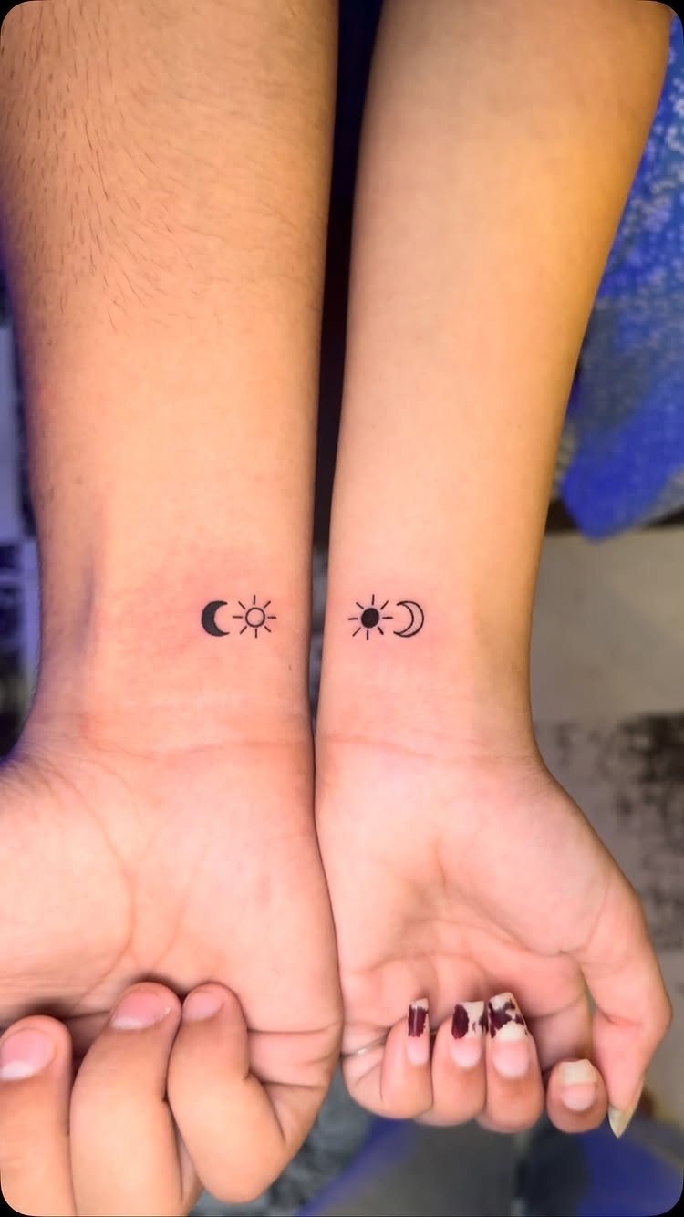 Two wrists with matching small black tattoos of a crescent moon and sun.
