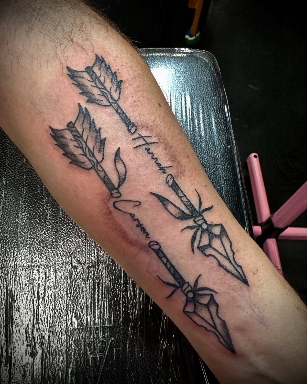 Two arrow tattoos with names on a person's forearm in black ink.