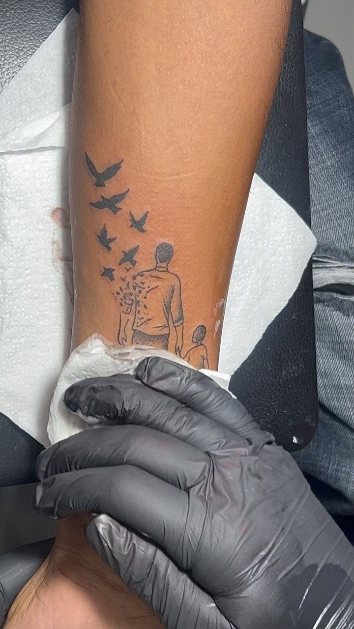 Tattoo on a forearm depicting a father and son with flying birds.