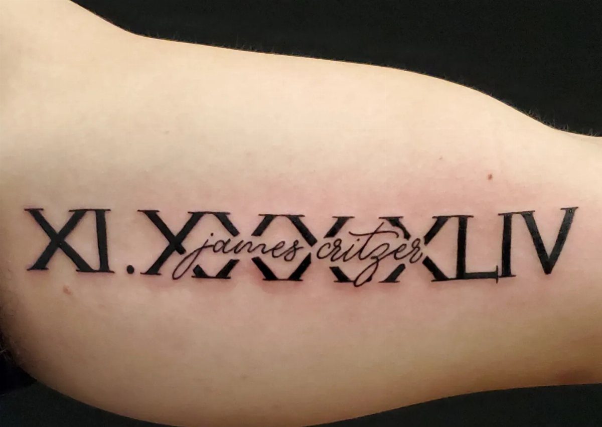 Roman numerals tattoo with a cursive name overlaid on a person's arm.