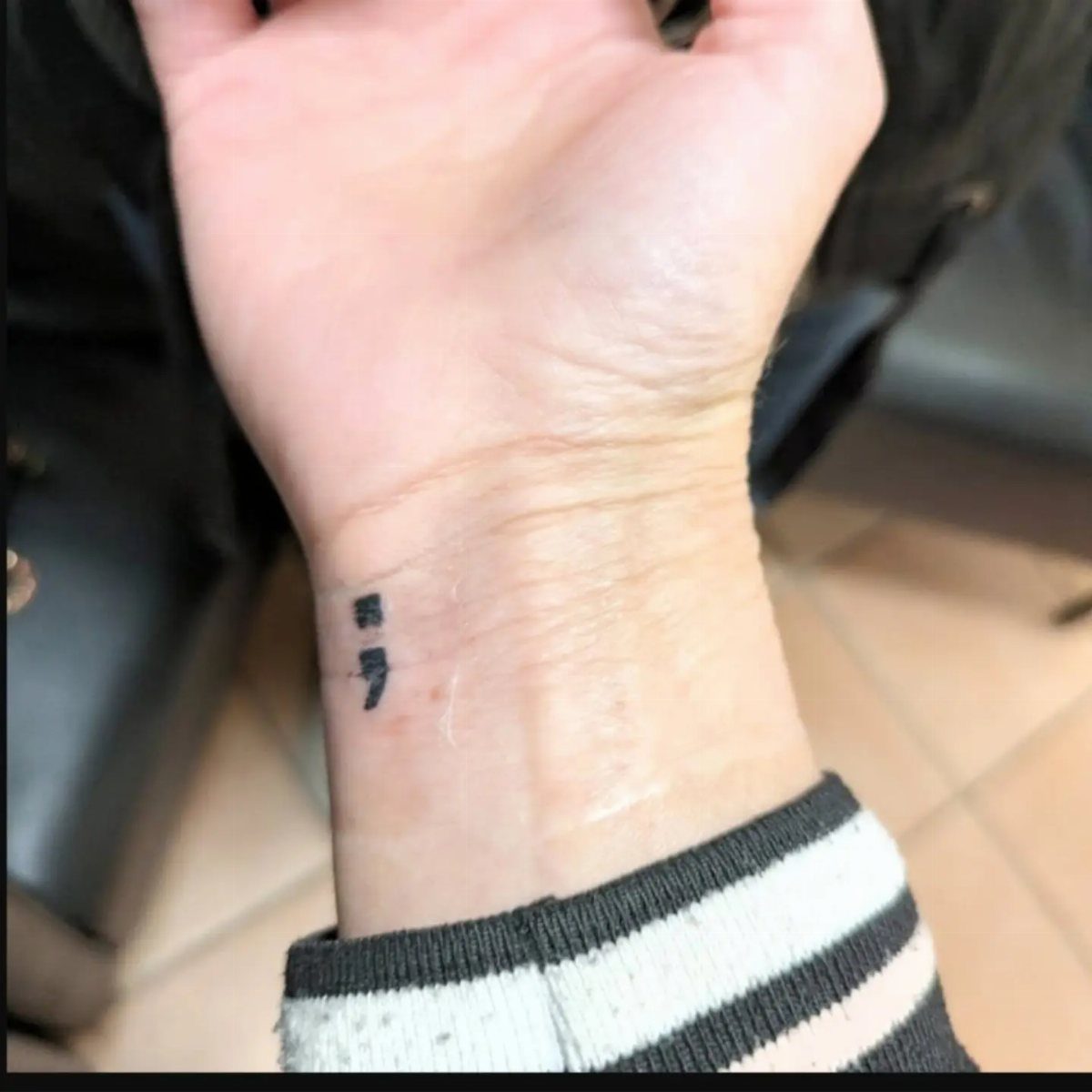Semicolon tattoo on a person's inner wrist with a striped sleeve visible.