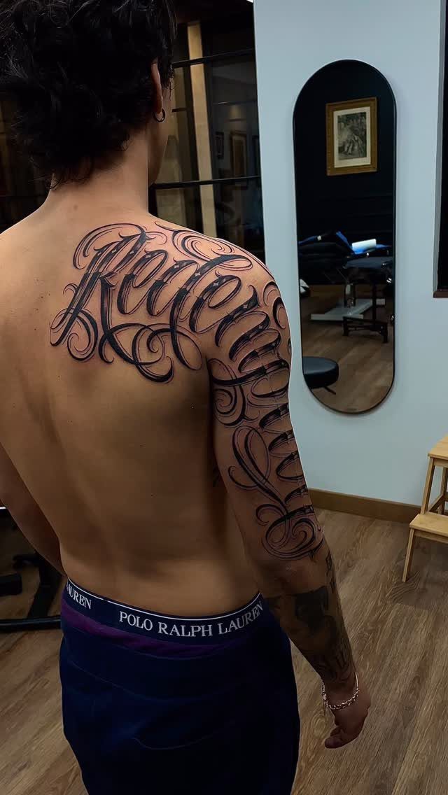 Ornate black and gray script tattoo on a man's back and upper arm.