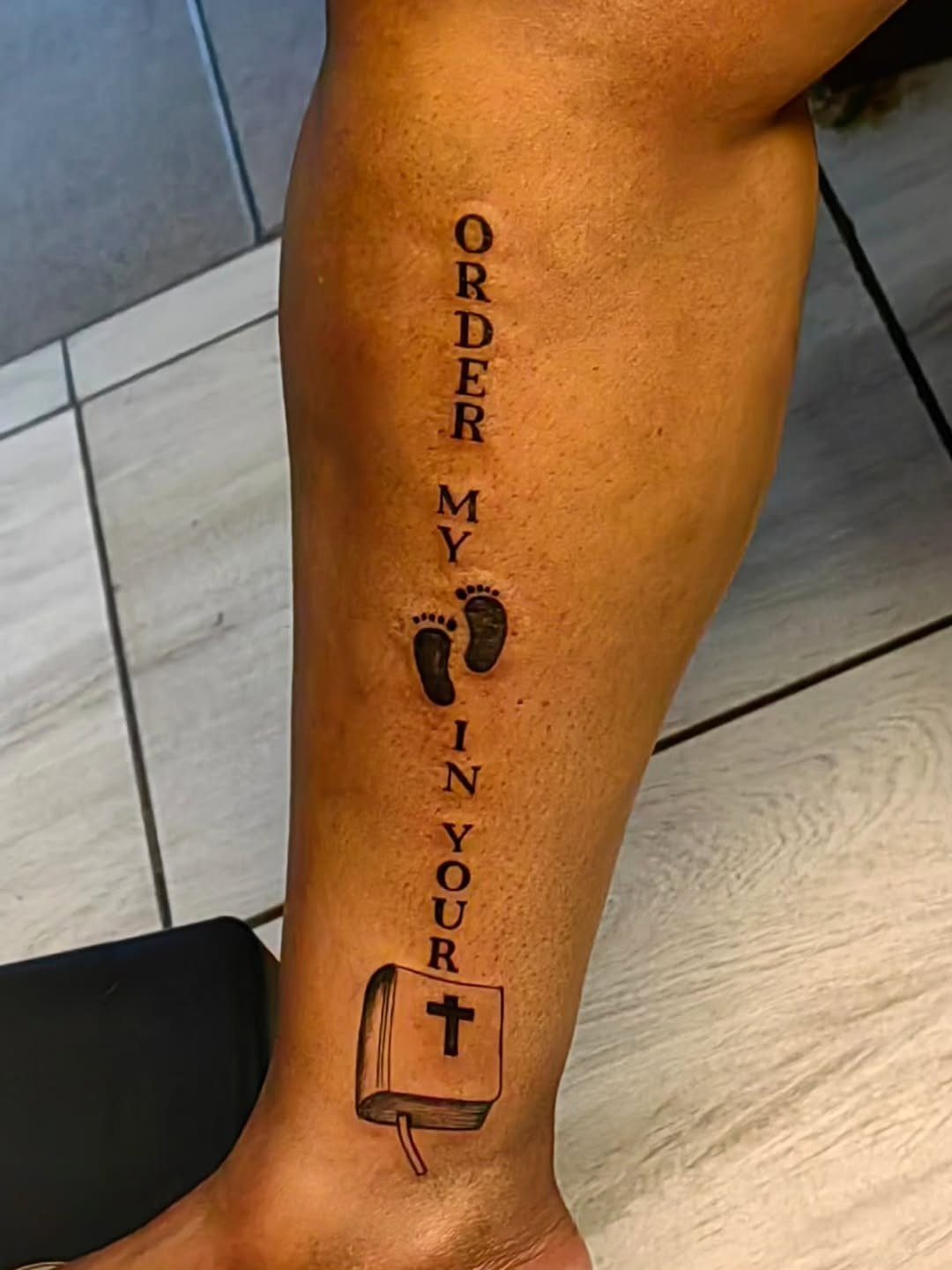 Tattoo on a lower leg featuring footprints, a Bible, and religious text.